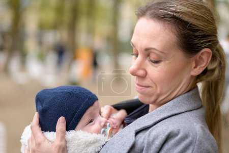 Photo for Portrait of an attractive blonde late-term mother in her 40s lovingly holding her newborn baby in a baby carrier and looking to him - Royalty Free Image