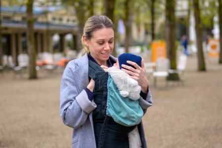 Téléchargez les photos : Jolie blonde mère à terme dans la quarantaine tenant amoureusement son nouveau-né dans un porte-bébé et regardant vers lui - en image libre de droit
