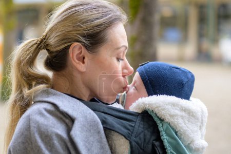 Foto de Vista lateral de una atractiva madre rubia de 40 años abrazando amorosamente a su bebé recién nacido en un portabebés y besándolo suavemente en la nariz - Imagen libre de derechos