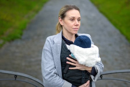 Téléchargez les photos : Femme épuisée tenant et portant son bébé dans un porte-bébé devant une rivière dans un parc - en image libre de droit