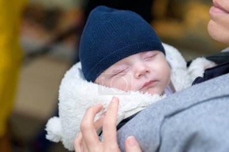 Téléchargez les photos : Gros plan d'un bébé dormant dans un porte-bébé avec sa mère portant une casquette bleue à l'extérieur - en image libre de droit