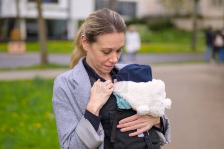 Glückliche Frau blickt auf ihr Baby, während sie es in einer Babytrage in einem Park hält und trägt