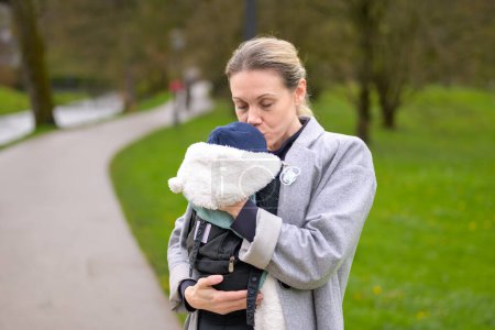 Téléchargez les photos : Femme heureuse embrassant son bébé qu'elle tient et porte dans un porte-bébé dans un parc - en image libre de droit