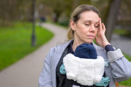 Photo for Close up of a exhausted and stressed woman holding and carrying her baby in a baby carrier in a park - Royalty Free Image