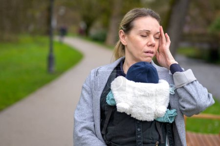 Photo for Exhausted and stressed woman holding and carrying her baby in a baby carrier in a park - Royalty Free Image