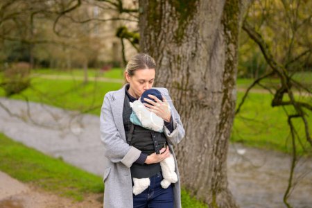 Téléchargez les photos : Femme heureuse embrassant son bébé qu'elle tient et porte dans un porte-bébé dans un parc - en image libre de droit