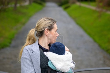 Foto de Mujer agotada sosteniendo y llevando a su bebé en un portabebés y mirando hacia un lado frente a un río en un parque - Imagen libre de derechos