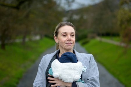 Photo for Happy woman holding and carrying her baby in a baby carrier in front of a river in a park - Royalty Free Image