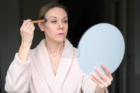 Téléchargez les photos : Femme blonde appliquant le maquillage avec un pinceau et regardant dans un miroir à la maison - en image libre de droit