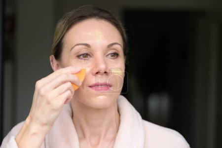 Téléchargez les photos : Femme blonde appliquant le maquillage avec une éponge à la maison - en image libre de droit