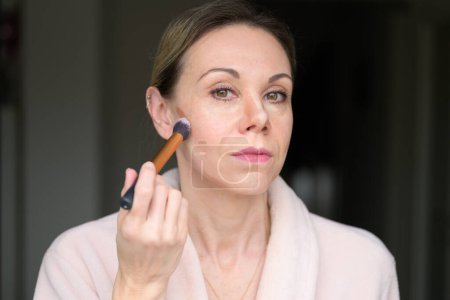 Téléchargez les photos : Femme blonde appliquant le maquillage avec un pinceau à la maison - en image libre de droit