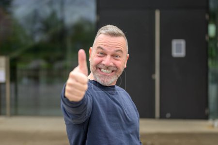 Téléchargez les photos : Enthousiaste motivé attrayant homme d'âge moyen donnant un pouce geste d'approbation et de succès avec un sourire rayonnant - en image libre de droit