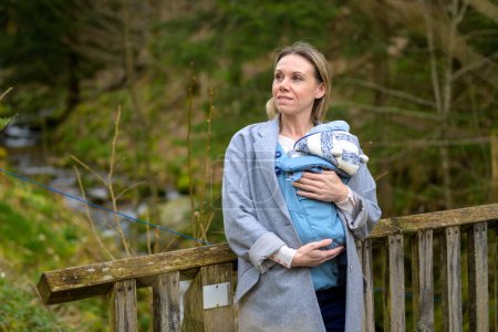 Téléchargez les photos : Femme heureuse regardant de côté tout en le tenant et le portant dans un porte-bébé debout sur un pont dans un parc - en image libre de droit