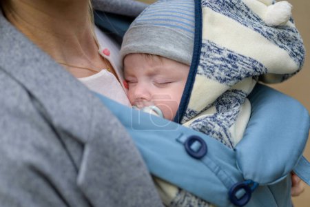 Téléchargez les photos : Gros plan d'un bébé dormant dans un porte-bébé sur la poitrine ou le décolleté de sa mère, dans une intimité exceptionnelle portant un chapeau rayé bleu et blanc - en image libre de droit