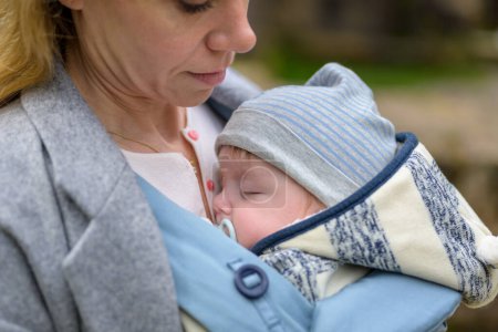 Téléchargez les photos : Gros plan d'un bébé dormant dans un porte-bébé sur la poitrine ou le décolleté de sa mère, dans une intimité exceptionnelle portant un chapeau rayé bleu et blanc - en image libre de droit