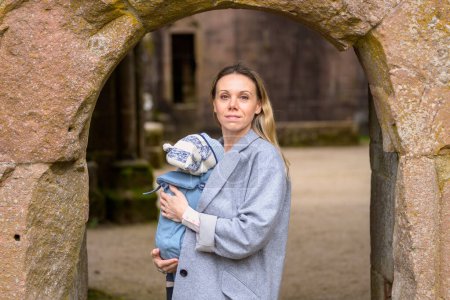 Téléchargez les photos : Portrait d'une femme heureuse regardant vers la caméra tout en tenant et en portant son bébé dans un porte-bébé dans une ruine - en image libre de droit