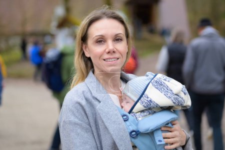 Téléchargez les photos : Portrait d'une femme heureuse regardant vers la caméra tout en le tenant et le portant dans un porte-bébé dans une ville - en image libre de droit