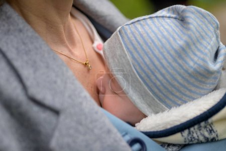 Téléchargez les photos : Gros plan d'un bébé dormant dans un porte-bébé, dans une extraordinaire proximité de sa mère portant un chapeau rayé bleu et blanc - en image libre de droit