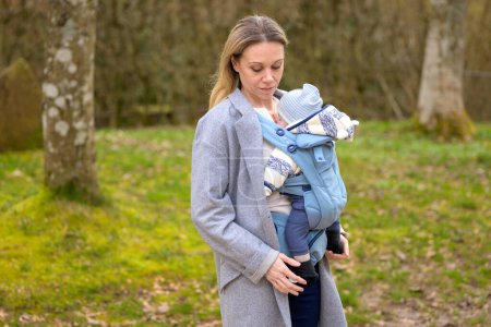 Téléchargez les photos : Femme heureuse regardant son bébé tout en le tenant et le portant dans un porte-bébé dans un parc - en image libre de droit