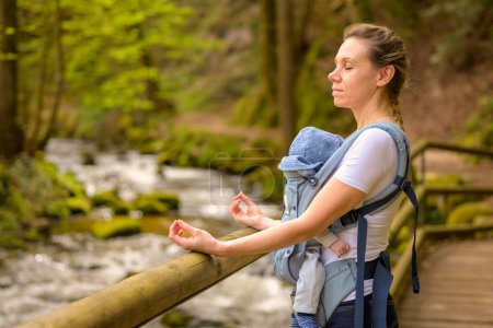 Téléchargez les photos : Femme heureuse avec les yeux fermés faire du yoga pendant un moment pour se calmer tout en tenant et en portant son bébé dans un porte-bébé dans un parc - en image libre de droit