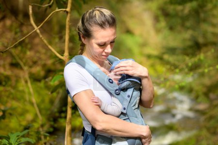 Téléchargez les photos : Femme heureuse regardant son bébé tout en le tenant et le portant dans un porte-bébé - en image libre de droit