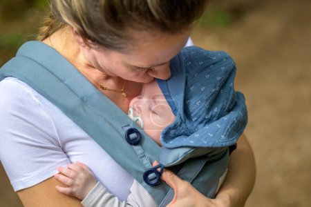 Téléchargez les photos : Extrême gros plan d'un bébé dormant dans un porte-bébé sur la poitrine ou le décolleté de sa mère, dans une intimité exceptionnelle portant un chapeau bleu - en image libre de droit