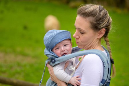 Glückliche Frau blickt auf ihr weinendes Baby, während sie es in einer Babytrage in einem Park hält und trägt
