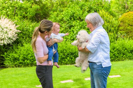 A heartwarming scene of three generations bonding in a beautiful garden. The young mother holds her baby, who excitedly reaches out towards a fluffy white dog held by the smiling grandmother.