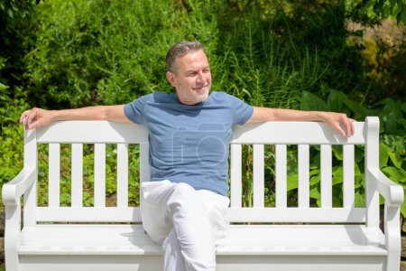 A mature man in casual attire sits on a white bench, reflecting and enjoying the peaceful surroundings of a lush park.
