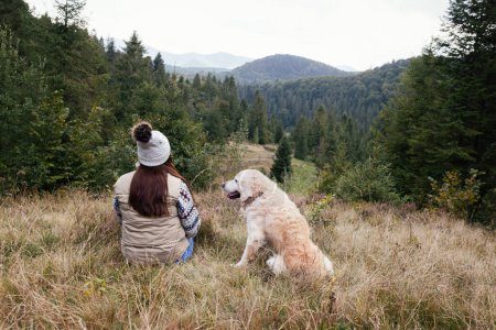 Photo for Young woman in a bobble hat and grey jacket with a golden retriever dog in the mountains. Autumn mood. Traveling with a pet. Rewilding outdoors journey concept. - Royalty Free Image