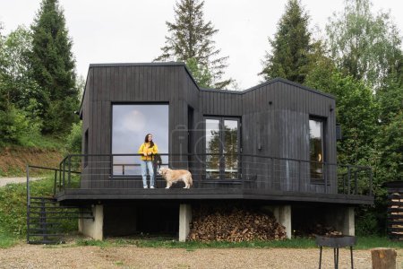 Woman wearing yellow coat and blue jeans and golden retriever dog near modern scandinavian style wooden cabin. Slow living and hygge lifestyle concept.