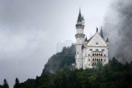 Photo for Castle of Neuschwanstein in Fussen, stunning neo gothic palace of the XIX century and most famous landmark of Bavaria, Germany. View from afar on a rainy misty day - Royalty Free Image
