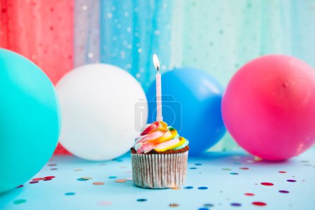 Foto de Colorido cupcake con una vela y globos de aire sobre fondo azul, tarjeta de aniversario - Imagen libre de derechos