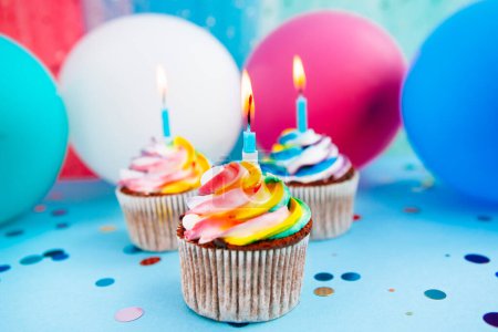 Foto de Tres cupcake de color arco iris con velas sobre fondo azul con globos de aire de colores, tarjeta de celebración - Imagen libre de derechos