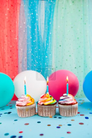 Foto de Tres cupcake de color arco iris con velas sobre fondo azul con globos de aire, tarjeta de celebración vertical - Imagen libre de derechos
