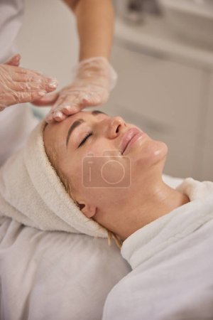 Gesichtsbehandlung, Ästhetische Dienstleistungen, Hautpflege-Beratung. Fachkundige Kosmetikerin massiert entspannt weibliche Klientin sanft während beruhigender Gesichtsbehandlung in sauberer und heller klinischer Umgebung.