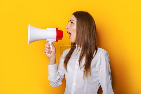 Photo for Young woman in a white shirt shouts into a megaphone on a yellow background. Concept for hiring, listing, help wanted. Banner. - Royalty Free Image
