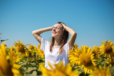 Photo for Young woman in a white t-shirt and glasses takes sunbathing on a sunflower field on a summer sunny day. - Royalty Free Image