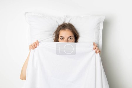 Young woman covering herself with a white sheet while lying on the bed. Top view, flat lay.