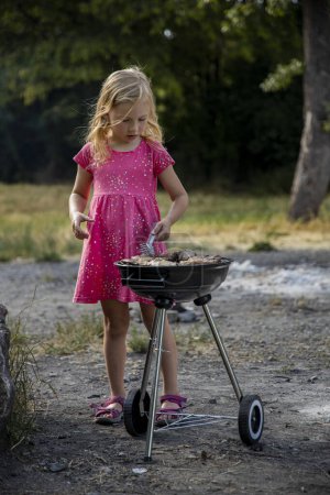 Téléchargez les photos : Petite fille enfant prépare la viande de poulet barbecue en plein air dans le parc dans la nature. - en image libre de droit
