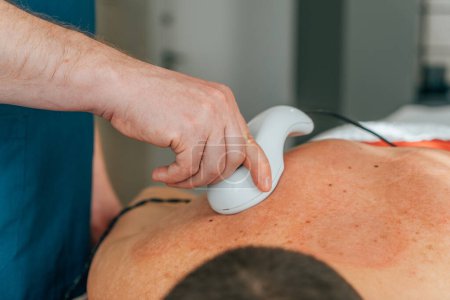 Nahaufnahme einer Physiotherapeutin, die während einer Therapiesitzung einen Perkussionsmassager auf den Rücken eines männlichen Patienten anwendet