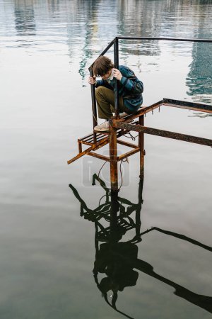 Thoughtful young individual man rests on rusty dock structure looking at his reflection into calm sea water, in serene natural setting.