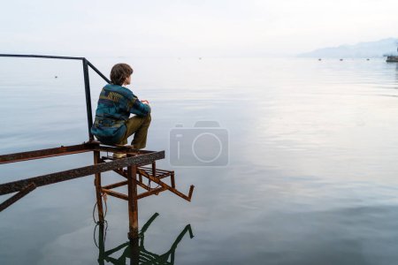 Foto de Hombre pensativo sentado solo en el muelle resistido por el mar tranquilo al atardecer, horizonte que se extiende más allá. - Imagen libre de derechos