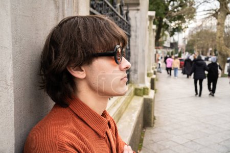 Ein junger Mann in orangefarbener Vintage-Jacke im 90er-Jahre-Stil und Vintage-Brille steht nachdenklich neben einer Säule auf der Stadtstraße. Modernes Retro- und Nostalgie-Trendkonzept.