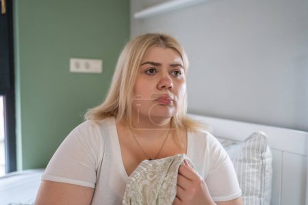 Concerned plus-size woman holding blanket, looks tired in her bedroom, problems with mental health.