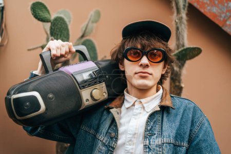 Junger Mann im Vintage-Stil hält eine tragbare Stereoanlage mit Kakteen im Hintergrund.