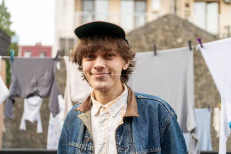 Smiling young man in denim jacket standing in front of hanging laundry.