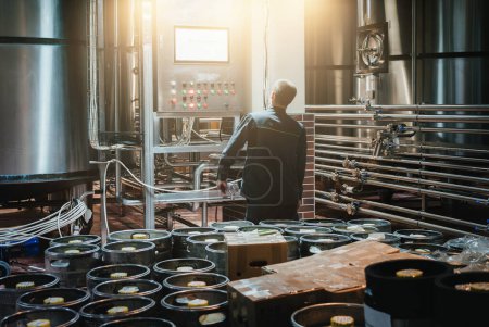 Braumeister überwacht Bierproduktion in Craft-Brauerei zwischen Fässern und glänzenden Edelstahlfässern