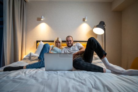 Paar liegt auf dem Bett, teilt Moment der Verbindung über Laptop-Bildschirm, in gemütlicher Schlafzimmeratmosphäre. 