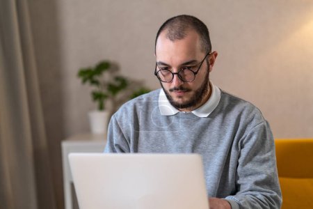 Porträt eines Mannes mit Brille, der zu Hause an seinem Laptop arbeitet.